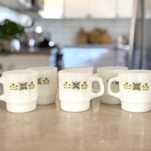 May include: A set of five white milk glass mugs featuring a repeating pattern of small green and brown floral and geometric motifs. Each mug has a cylindrical body with a stepped base and a D-shaped handle, designed for stacking.