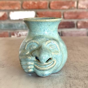 May include: A turquoise ceramic vase with a smiling face design. The vase has a rounded body with a wide opening and a detailed face sculpted on the front, including a nose, eyes, and a smiling mouth.