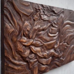 May include: A rectangular, dark brown wooden wall art piece with a carved, swirling design. The artwork has a textured surface and is mounted on a light gray wall. The wood grain is visible.