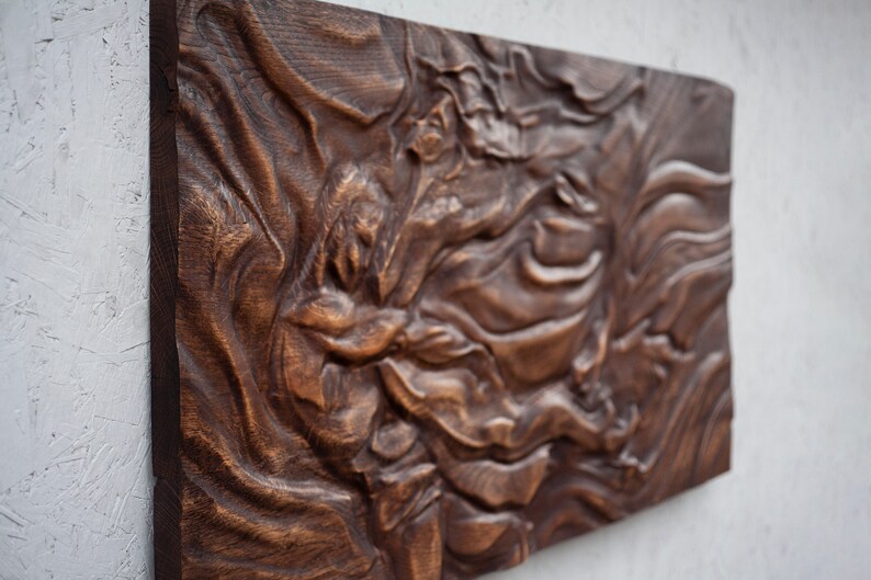 May include: A rectangular, dark brown wooden wall art piece with a carved, swirling design. The artwork has a textured surface and is mounted on a light gray wall. The wood grain is visible.