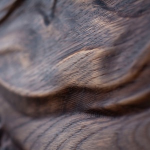May include: Close-up of a dark brown wooden surface with a wavy, textured pattern. The wood grain is visible, with lighter brown highlights contrasting against the darker tones. The image focuses on the natural texture and color variations.