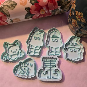 May include: A collection of light blue plastic cookie cutters in various Halloween shapes, including a bat, pumpkin, ghost, and witch. The cutters are arranged on a pink surface, with floral rolling pins in the background.