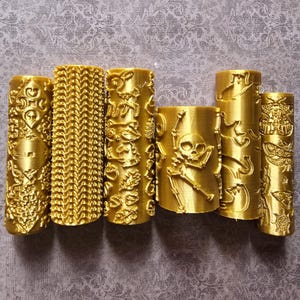 May include: Six gold-colored cylindrical objects with intricate embossed designs. The designs include a skull and crossbones, floral patterns, and other ornate details. The objects are arranged in a row against a patterned background.