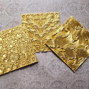 May include: Three gold-colored, square decorative panels with intricate embossed patterns. One panel features a floral design, another has a wave pattern, and the third has a detailed, textured design. The panels are displayed on a patterned gray surface.