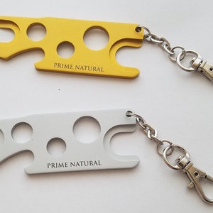 May include: Two keychains with a multi-tool design. The keychain on the top is gold and the keychain on the bottom is silver. Both keychains have the text "PRIME NATURAL" printed on them.
