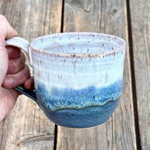 Peut inclure: Une tasse en céramique avec un émail bicolore, présentant une partie supérieure blanche tachetée et une partie inférieure bleu profond. La tasse possède un corps large et arrondi ainsi qu'une anse robuste, avec un motif de coulure distinct.