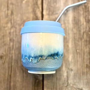 Peut inclure: Un mug de voyage en céramique avec un couvercle en silicone bleu clair et une paille en métal. Le mug présente un émail réactif avec une partie supérieure blanche qui se transforme en un motif goutte bleu profond sur une base grise mouchetée.