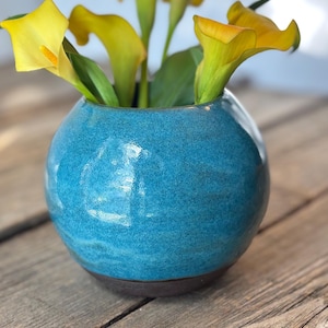 May include: A round, turquoise ceramic vase with a glossy, textured finish and a dark brown base. The vase contains several bright yellow calla lily flowers with green stems and leaves, creating a vibrant contrast between the blue and yellow.