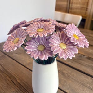 Peut inclure: Un bouquet de fleurs de marguerite en céramique de couleur lavande avec des centres jaunes, chacune fixée à une fine tige métallique verte. Les fleurs sont disposées dans un vase en céramique blanc mat, lisse, de forme arrondie et effilée.