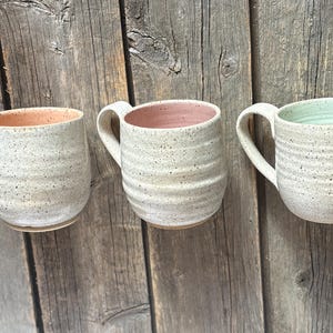 May include: Three ceramic mugs with a speckled off-white exterior and horizontal ribbed texture. Each mug features a distinct interior colour: one is light orange, one is dusty pink, and one is a pale mint green. Each has a curved handle.