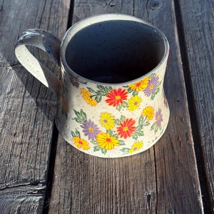 May include: A speckled beige ceramic mug featuring a floral design with yellow, red, and purple flowers. The mug has a curved handle and sits on a rustic wooden surface. The design highlights the handmade nature of the mug.