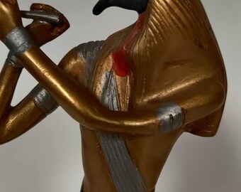 May include: A gold-colored statue of an Egyptian god with a black bird head and a white and gold headdress. The statue is holding a staff in its right hand.