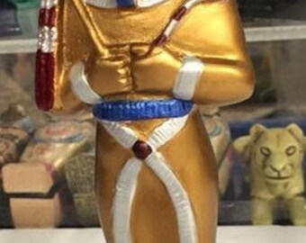 Peut inclure: Statue dorée d'un pharaon égyptien tenant un bâton. Le pharaon porte un couvre-chef bleu et blanc et une robe dorée et blanche. La statue est posée sur une base noire.
