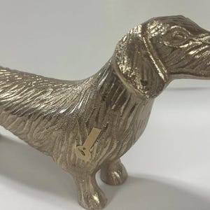 Pode incluir: Uma estatueta de cão dachshund de cor dourada metálica. O cão está em pé com a cabeça para cima e a cauda para baixo. A estatueta tem uma textura de pêlo detalhada e é um item decorativo.