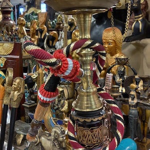 May include: An assortment of Egyptian-themed decorative items, including a hookah with a brown top and brass base, statues, and a turquoise hippo figurine. The hookah has red, black, and gold accents. Other items include gold statues and decorative canes.