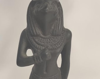 Peut inclure: Statue sombre d'une divinité égyptienne, peut-être Horus ou une figure similaire. La statue présente une tête d'oiseau, un collier détaillé et une jupe. La figure tient un objet dans sa main et se tient sur une base rectangulaire.