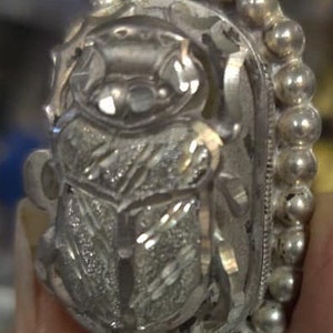May include: A silver ring with a detailed design of a scarab beetle. The ring is adorned with a beaded border.