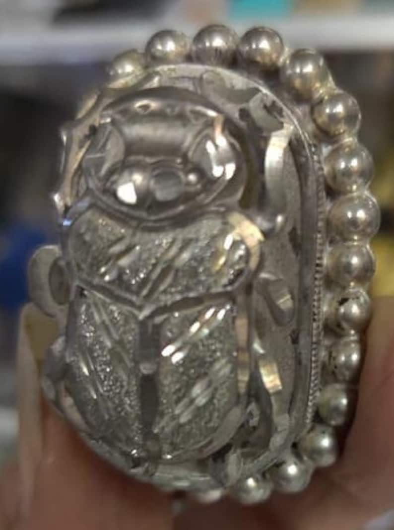 May include: A silver ring with a detailed design of a scarab beetle. The ring is adorned with a beaded border.