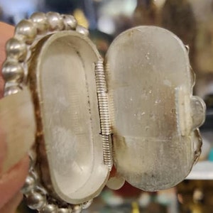 May include: A silver oval-shaped hinged box with a beaded edge. The box is open, revealing a spring hinge.