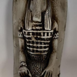 Peut inclure: Une statue en céramique blanche d'un dieu égyptien assis sur un trône. Le dieu a une tête de chacal et porte un vêtement rayé.