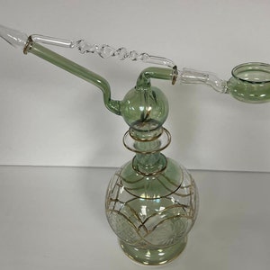 May include: A green glass hookah with a gold trim and a clear glass stem. The hookah has a round base with a decorative pattern and a small bowl on the top.