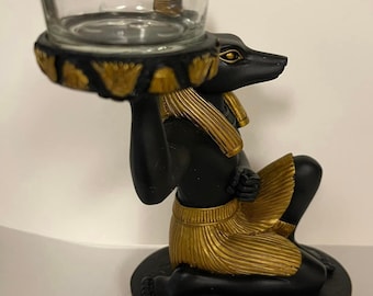 May include: Black and gold Anubis candle holder with a glass votive. The base of the holder features hieroglyphics.