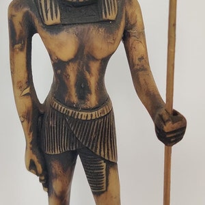 May include: A tan-colored statue of a standing figure with a lion's head, holding a staff with a gold-colored top. The figure is wearing a skirt and has detailed facial features. The statue is on a rectangular base.