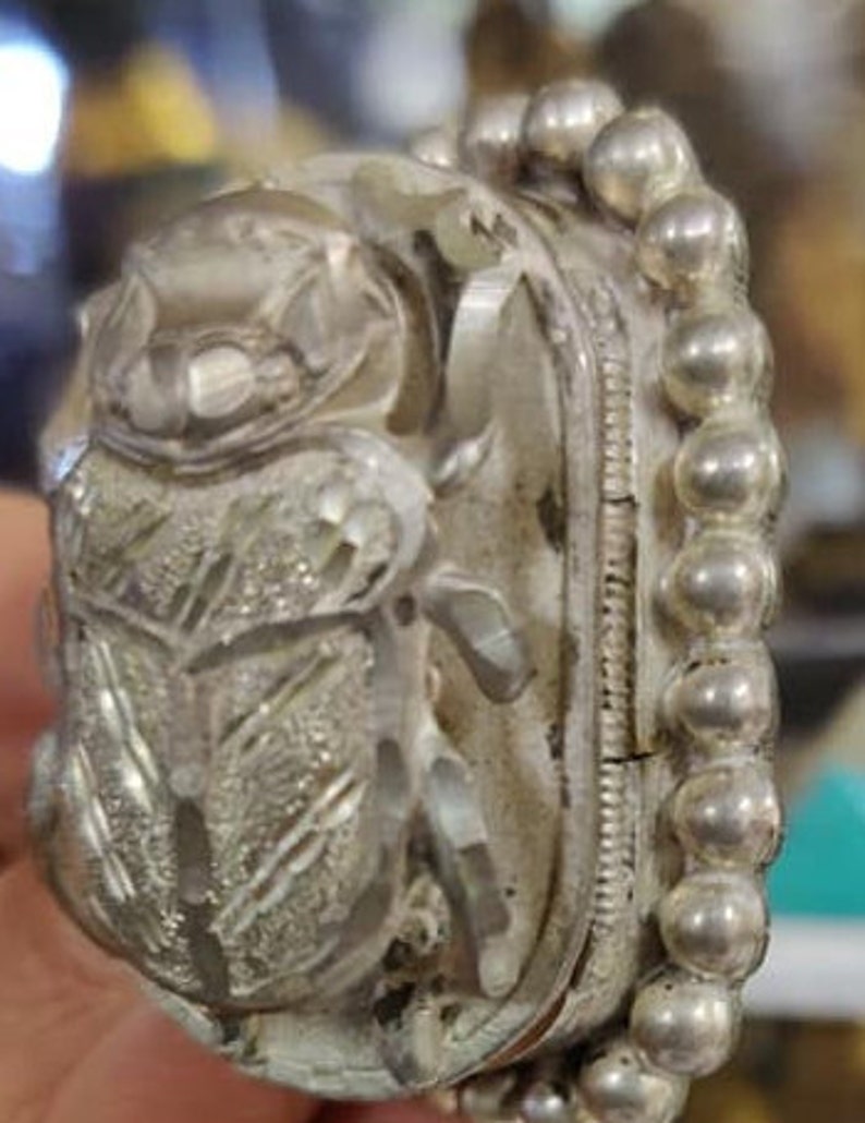 May include: A silver ring with a detailed beetle design. The beetle is made of clear crystal and is surrounded by a beaded band.