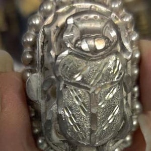 May include: A silver ring with a detailed, engraved scarab beetle design. The ring is surrounded by a beaded border.