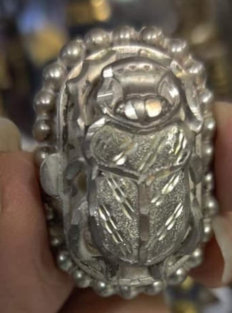 May include: A silver ring with a detailed, engraved scarab beetle design. The ring is surrounded by a beaded border.