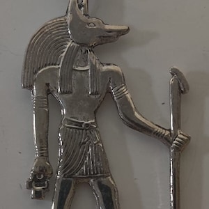 Op de afbeelding: Zilveren hanger met een Anubis-figuur met een staf. De hanger is gedetailleerd met ingewikkelde gravures en een lus om een ​​ketting aan te bevestigen.