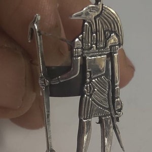 Puede incluir: Un anillo de plata que representa una deidad egipcia, posiblemente Thoth, con cabeza de pájaro y báculo. La figura está intrincadamente detallada. El anillo está sostenido por una mano, mostrando la artesanía.