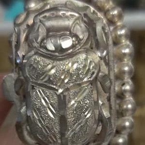 May include: A silver bracelet with a detailed beetle charm. The beetle is intricately carved and has a textured surface. The bracelet is made of small silver beads.
