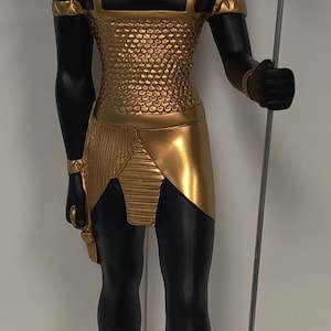 Puede incluir: Una estatua negra y dorada de un dios egipcio con cabeza de chacal, que lleva un tocado dorado y sostiene un bastón. La estatua está de pie sobre una base de madera con jeroglíficos.