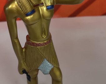 Peut inclure: Une figurine dorée d'un dieu égyptien à tête de chacal, portant une jupe et debout sur une base brune.
