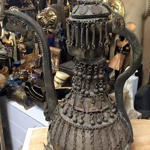 May include: An antique metal pitcher with a long, curved handle and ornate detailing. The pitcher is adorned with hanging coin-like charms and decorative metalwork. The overall color is a tarnished bronze, suggesting age and historical significance. The pitcher is likely a decorative item.