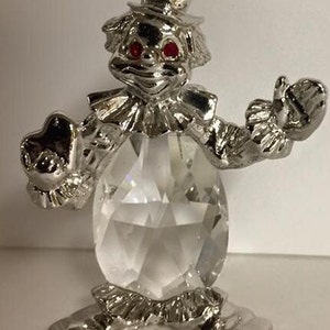 May include: A silver figurine of a clown with a crystal body. The clown is wearing a hat and has red eyes. The figurine is standing on a white surface.