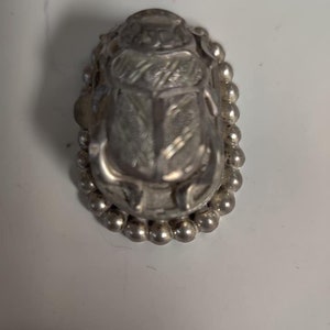 May include: A silver scarab beetle charm with a beaded border. The beetle is detailed with intricate lines and a raised design.