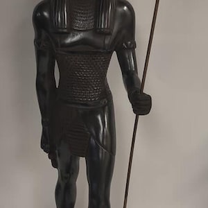 Peut inclure: Statue noire d'un dieu égyptien à tête de chacal, tenant un bâton surmonté d'un oiseau. La statue est posée sur une base noire avec des hiéroglyphes.
