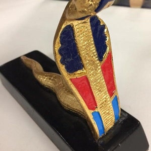 Könnte beinhalten: Eine goldfarbene Schlangenskulptur mit blauem und rotem Design auf ihrem Körper, montiert auf einem schwarzen rechteckigen Sockel. Der Kopf der Schlange ist erhoben, und ihr Körper krümmt sich entlang des Sockels. Die Skulptur hat ein strukturiertes Aussehen.