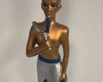 Peut inclure: Une statue dorée d'un pharaon égyptien portant une couronne rouge et blanche, tenant un bâton dans sa main droite. La statue est posée sur une base en bois.