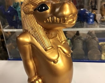 Peut inclure: Une statue dorée d'un hippopotame à tête humaine, portant un couvre-chef noir. La statue est posée sur une base avec des hiéroglyphes.