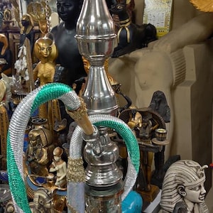 May include: A silver hookah with a glass base and a decorative green and silver hose. The hookah has a metal stem and a bowl at the top. The background includes various Egyptian-themed statues.
