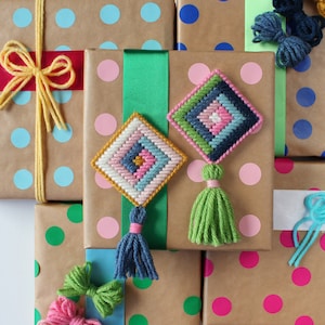 Peut inclure: Plusieurs cadeaux emballés avec du papier brun, des pois colorés et des rubans. Deux ornements en forme de losange, cousus à la main, avec des glands sont présentés. Les cadeaux sont ornés de nœuds et de fil de différentes couleurs, créant une esthétique festive et artisanale.