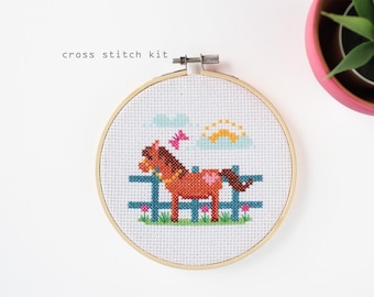 May include: A cross stitch kit featuring a brown horse with a pink heart on its side, standing behind a blue fence. The design includes a butterfly, clouds, and a sun. The text "cross stitch kit" is above the design.