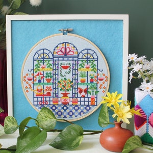 May include: A framed cross-stitch embroidery depicting a colorful indoor garden scene with plants, flowers, and a birdhouse. The embroidery is in a round hoop and is surrounded by a light blue background.