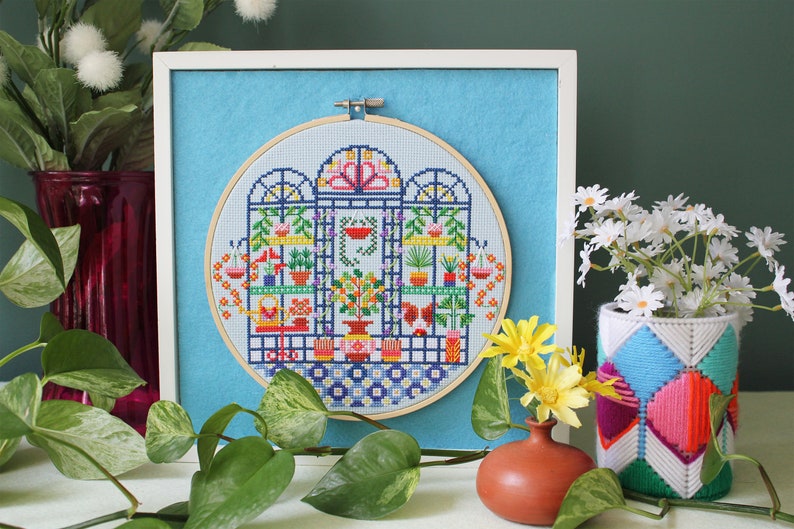 May include: A framed cross-stitch embroidery depicting a colorful indoor garden scene with plants, flowers, and a birdhouse. The embroidery is in a round hoop and is surrounded by a light blue background.
