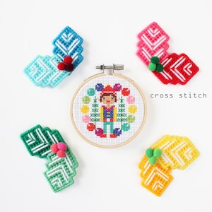Peut inclure: Un kit de point de croix avec un cercle de broderie terminé représentant une personne entourée de fleurs colorées. Quatre ensembles d'éléments décoratifs cousus assortis en bleu, rouge, vert et jaune sont également inclus. Le texte "cross stitch kit" est visible.