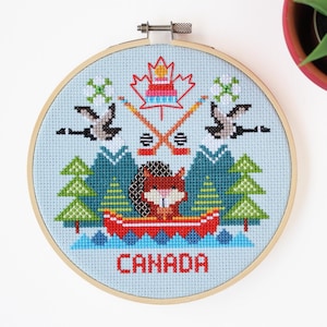 May include: Cross-stitch embroidery in a wooden hoop featuring a Canadian theme. The design includes a red canoe with a fox, mountains, trees, hockey sticks, geese, and the word "CANADA" in red letters. The background is light blue.