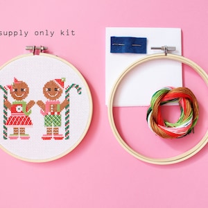 May include: A cross-stitch kit featuring two gingerbread people holding candy canes. The kit includes a white Aida cloth, embroidery floss, and a wooden embroidery hoop. The text "supply only kit" is printed on a white background.